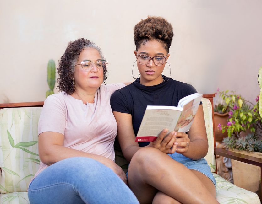 A mother and daughter sit closely together reading a book, sharing a quiet conversation.