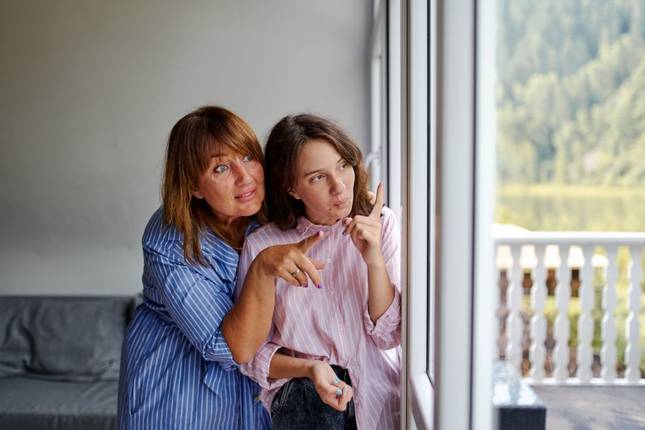 A mother and adult daughter share a quiet, reflective moment together, gazing out a window as if opening up about things left unsaid.