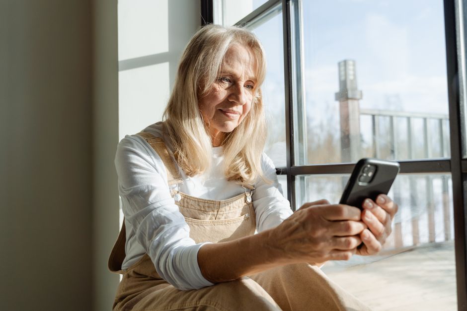 Senior woman checking her phone by the window before stepping outside, much like checking the weather before leaving home.
