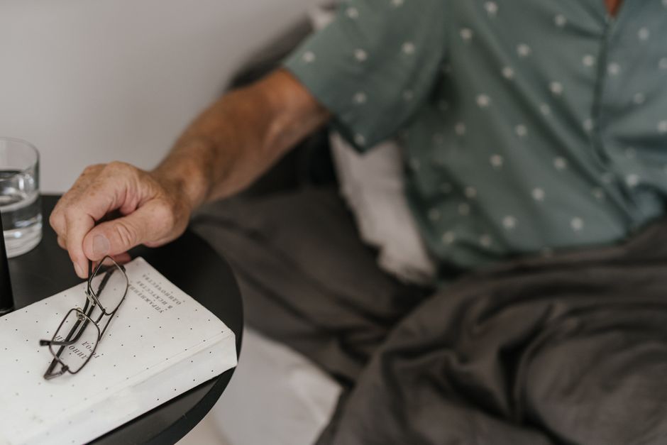 An elderly man sets his glasses down on a nightstand book as he settles into bed early for the night.