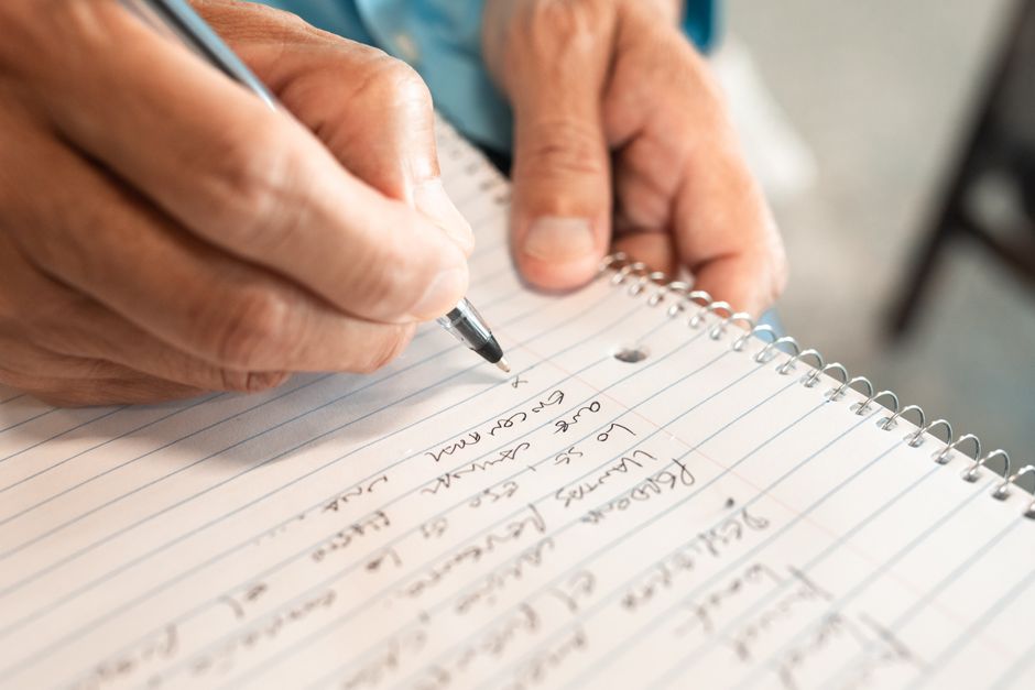 An older person's hands writing a list by hand in a spiral notebook, a habit that helps keep the list in their head
