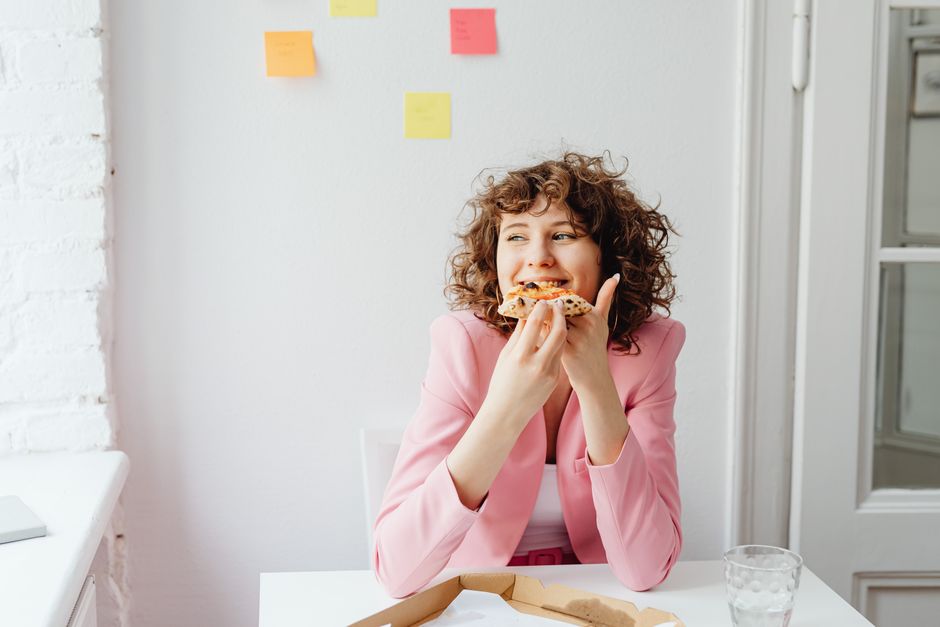 A woman taking a satisfying bite of pizza at her desk, proving that eating lunch is not optional.