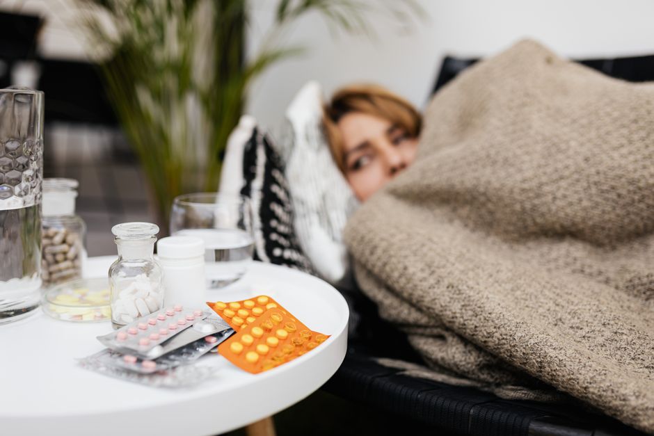 A person resting sick under a blanket next to a table full of pain relievers and medicine bottles.