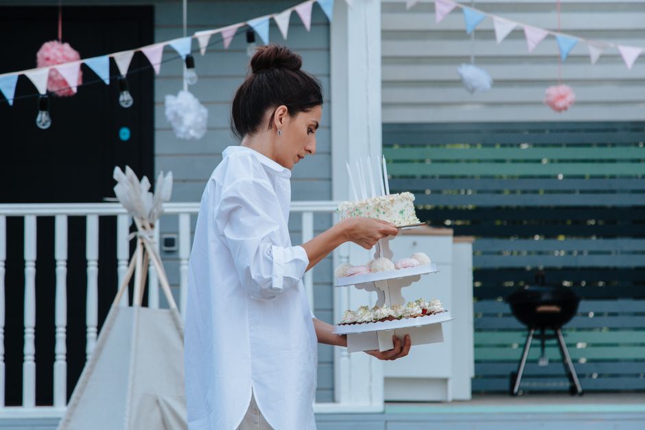 A woman wearily carries a birthday cake outside while setting up for a party she organized.