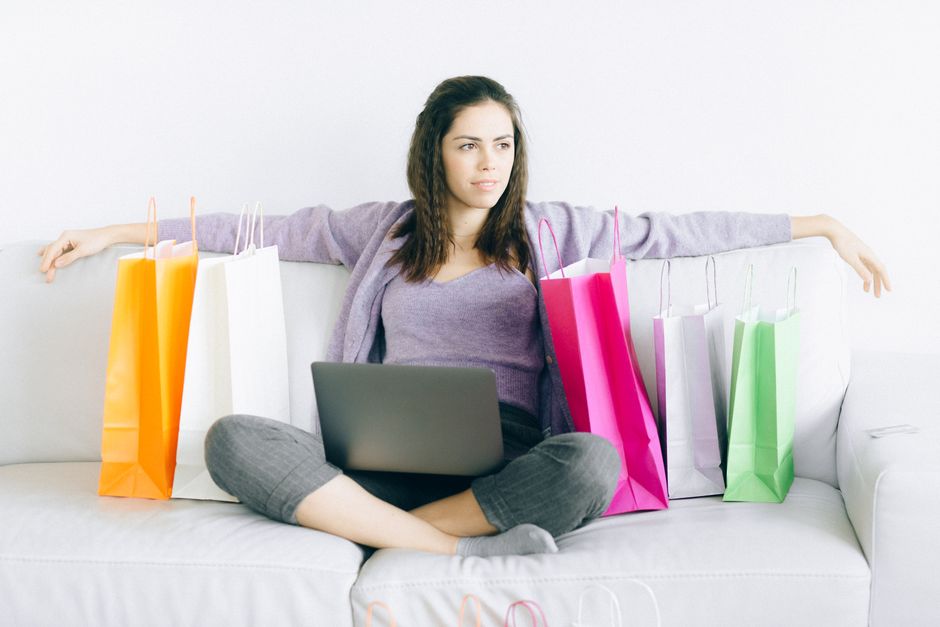 A woman surrounded by shopping bags looks unenthusiastic while browsing on her laptop, reflecting mixed feelings toward Black Friday shopping.