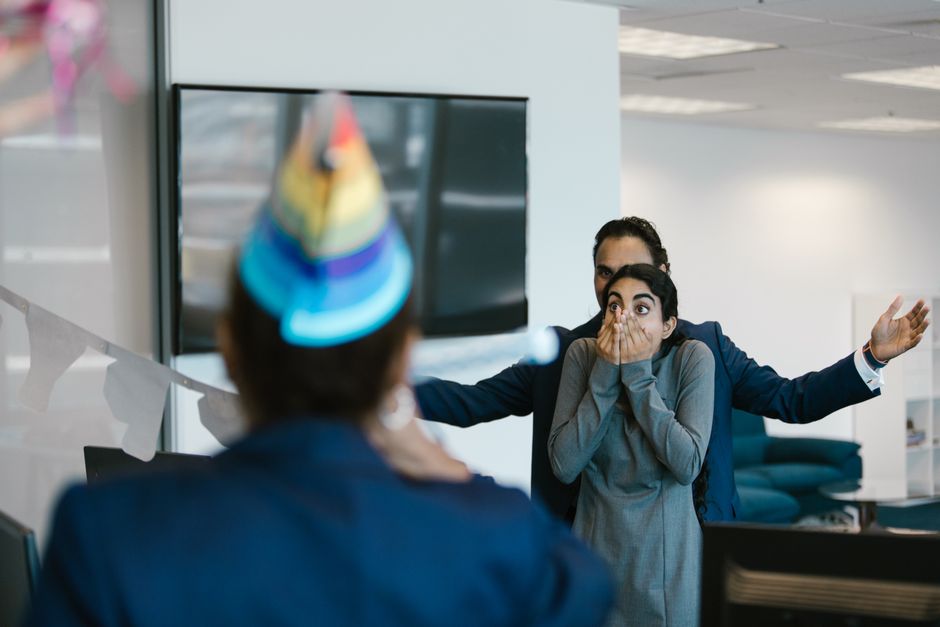A woman looks visibly stressed and overwhelmed as she's caught off guard by a surprise celebration at work.