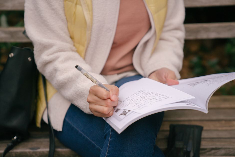 A woman writes with a pen in her notebook while her purse sits nearby on a bench